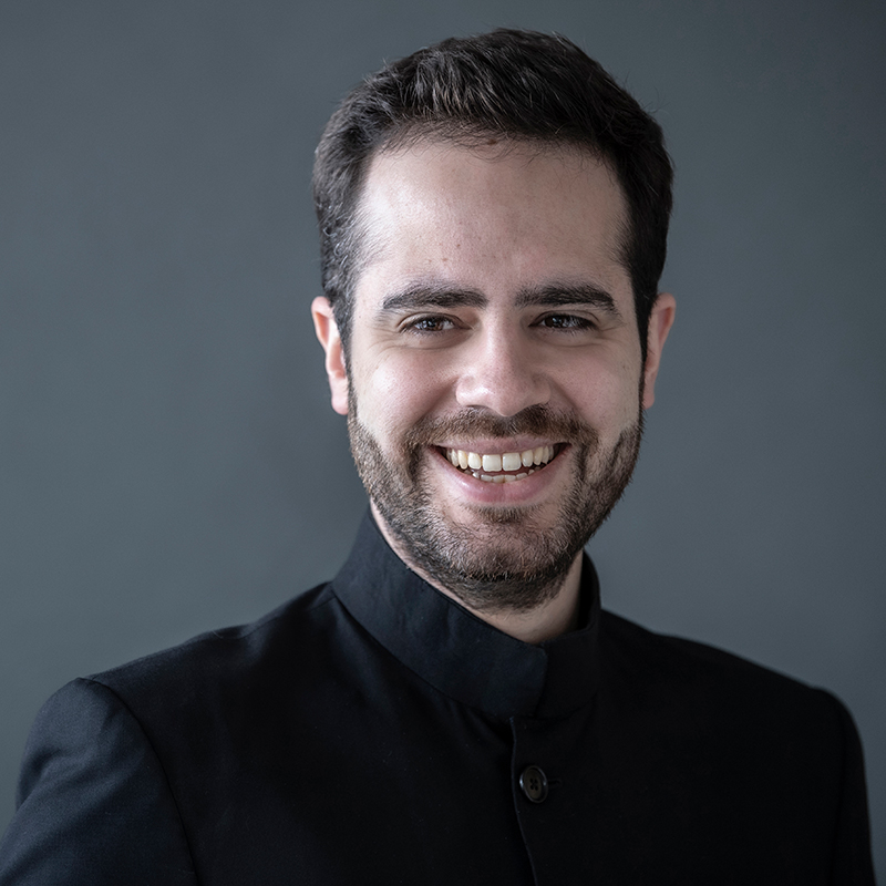 A man with short dark hair and a beard smiles at the camera. He is wearing a black jacket with a stand-up collar and is posed against a plain grey background.