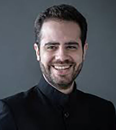 Dinis Sousa, a man with short dark hair and a beard is smiling. He is wearing a dark-coloured jacket with a high collar, and the background is plain and grey.