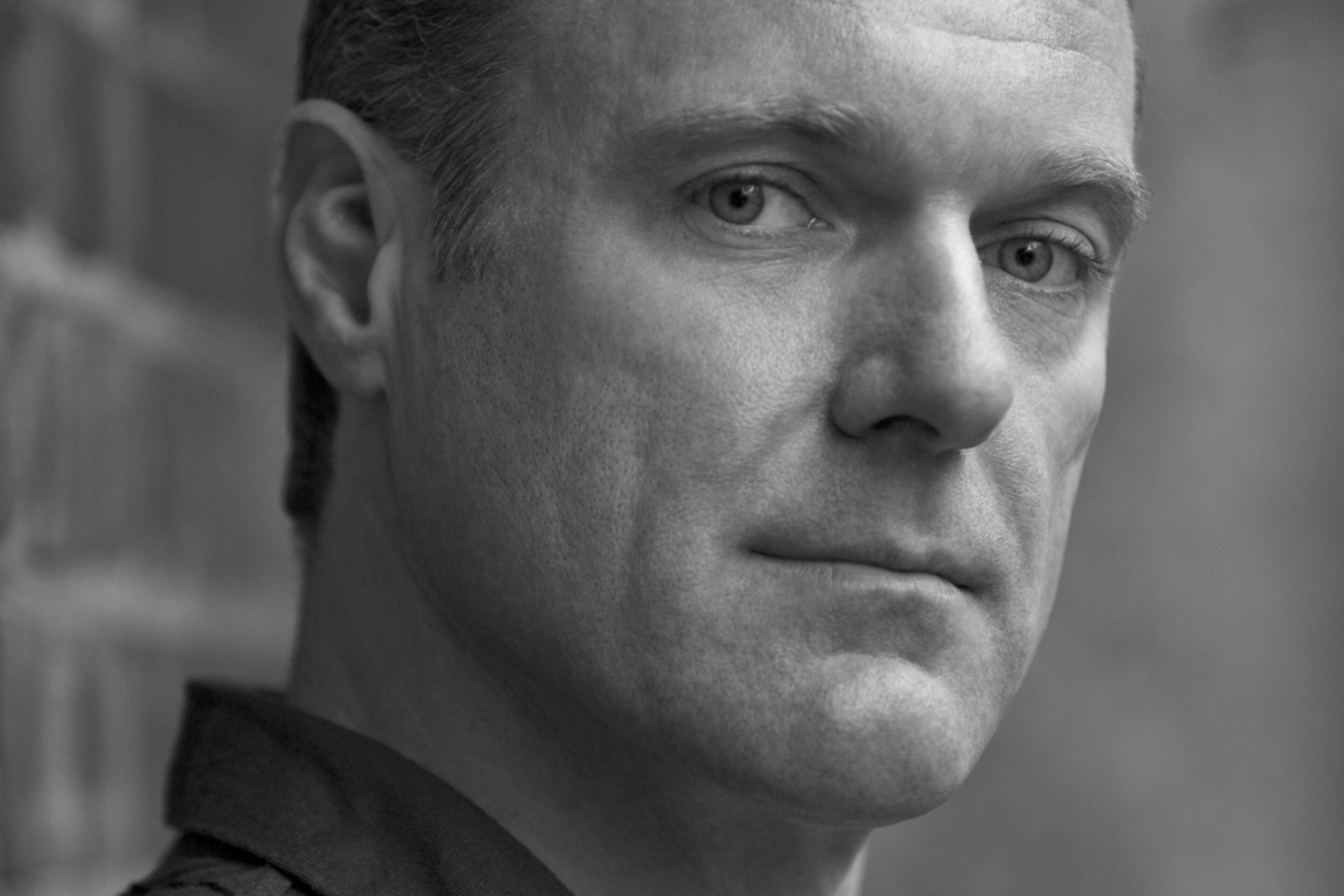 A man with short hair and a serious expression is looking slightly past the camera. He is wearing a dark collared shirt and standing next to a brick wall in an outdoor setting. The image is in black and white.