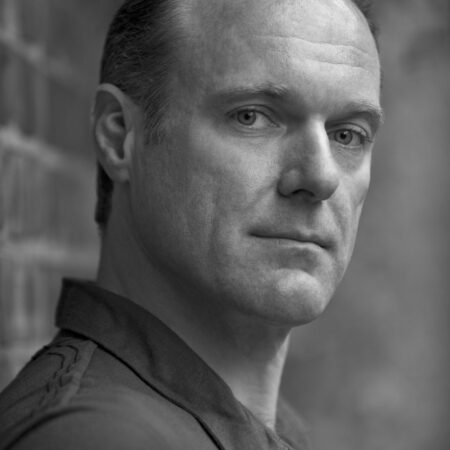 A man with short hair and a serious expression is looking slightly past the camera. He is wearing a dark collared shirt and standing next to a brick wall in an outdoor setting. The image is in black and white.