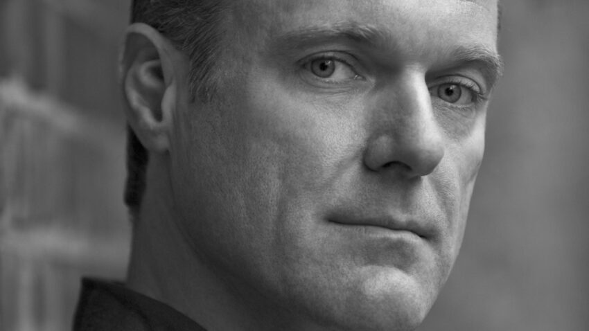 A man with short hair and a serious expression is looking slightly past the camera. He is wearing a dark collared shirt and standing next to a brick wall in an outdoor setting. The image is in black and white.