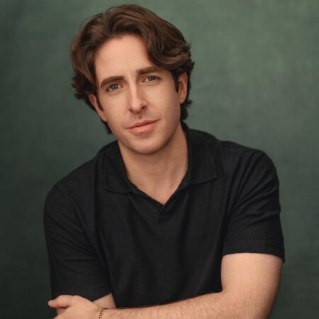 A young man with wavy brown hair and blue eyes, Elgan Llŷr Thomas sits against a green backdrop, wearing a short-sleeved black shirt and a gold bracelet, his arms gently crossed and his expression relaxed.