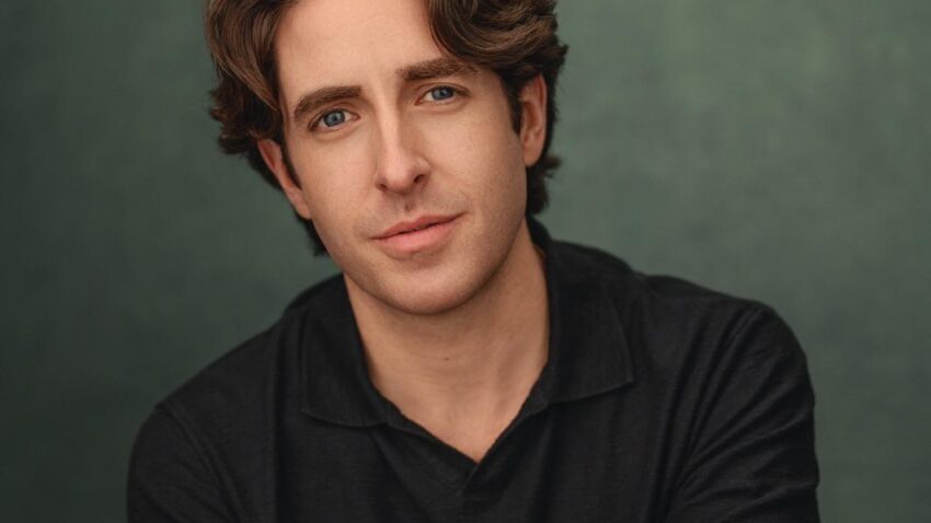 A young man with wavy brown hair and blue eyes, Elgan Llŷr Thomas sits against a green backdrop, wearing a short-sleeved black shirt and a gold bracelet, his arms gently crossed and his expression relaxed.