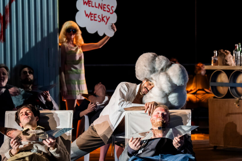 A woman in sunglasses holds a sign reading WELLNESS WEEKSY behind people lounging on chairs, one with a large white wig shaped like clouds. The playful, theatrical scene evokes the spirited world of the Mahagonny opera.