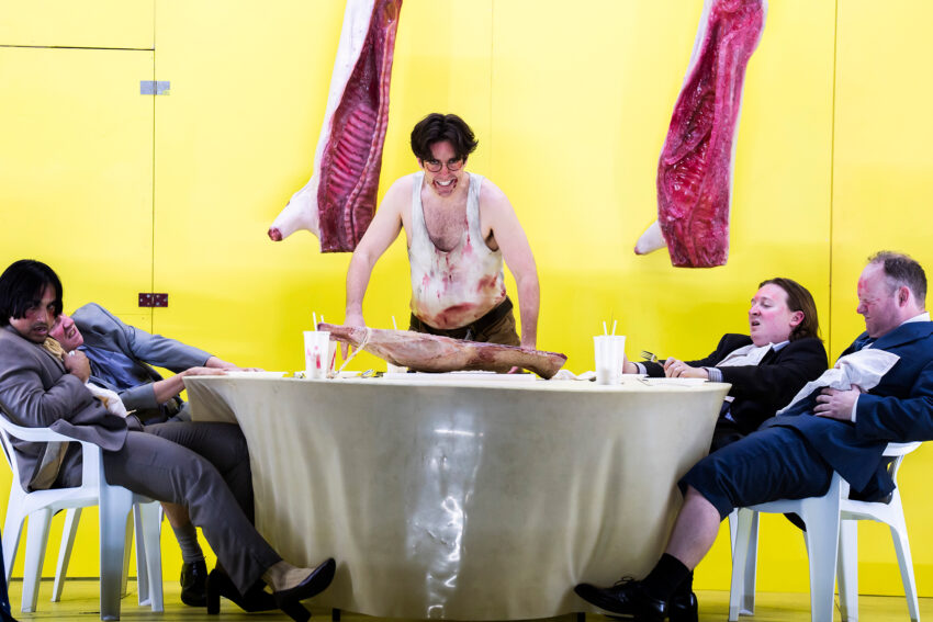 A man in a dirty vest stands behind a table with a large slab of meat, while three men in suits slump in white plastic chairs around him—a scene evoking the gritty satire of Bertolt Brecht’s Mahagonny opera. The background is bright yellow with hanging cuts of meat.