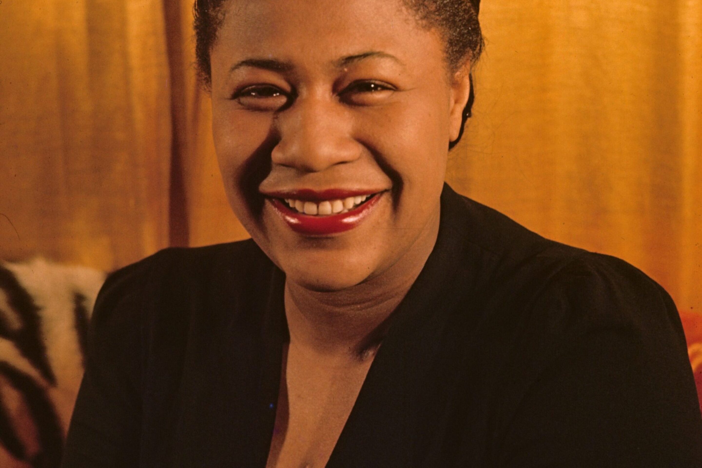 A smiling woman with dark curly hair wearing a black dress with gold embroidery sits in front of a gold curtain, evoking the timeless elegance of "Summertime" by George Gershwin.