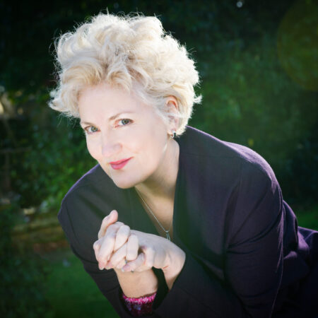 A woman with short, curly blonde hair wearing a dark jacket leans forward and smiles at the camera, with hands clasped. The background is outdoors with greenery and sunlight.