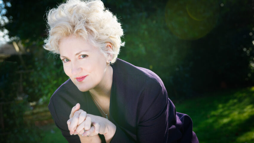 A woman with short, curly blonde hair wearing a dark jacket leans forward and smiles at the camera, with hands clasped. The background is outdoors with greenery and sunlight.