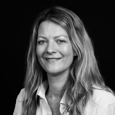 A woman with long, wavy hair is smiling at the camera. She is wearing a light-coloured button-up shirt and is set against a plain black background. The image is in black and white.