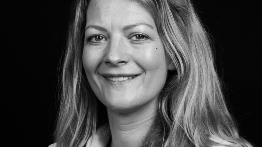 A woman with long, wavy hair is smiling at the camera. She is wearing a light-coloured button-up shirt and is set against a plain black background. The image is in black and white.