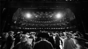 A group of people wearing hats stand on a theatre stage under bright lights, facing an illuminated audience seating area with balconies, during a black and white 2026 Gilbert & Sullivan Sing-Along event.