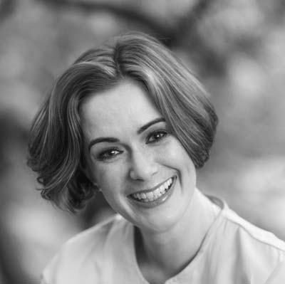 Harriet Eyley, a woman with short, wavy hair, smiles warmly at the camera. The image is in black and white, with a softly blurred outdoor background.