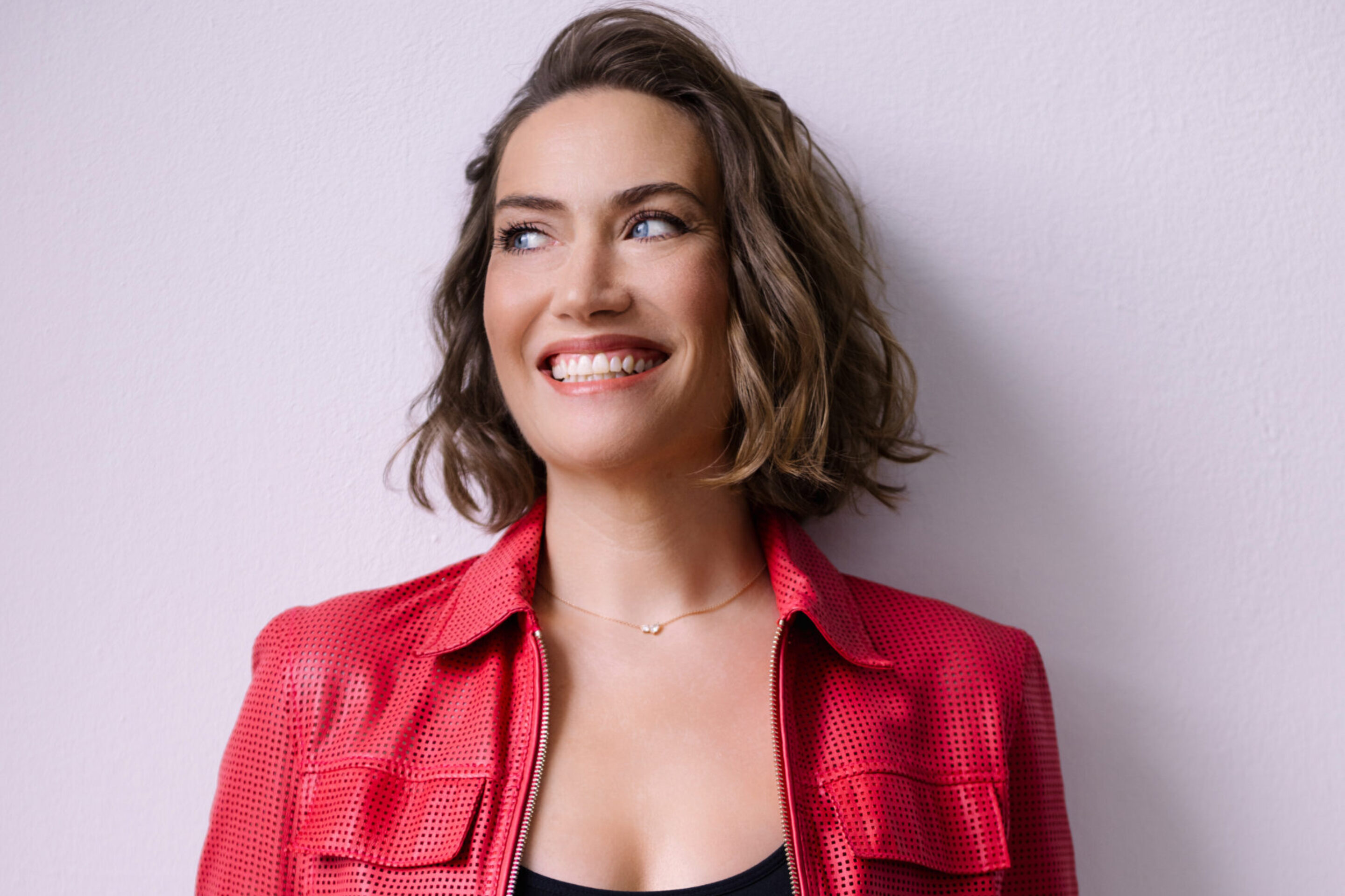 Opera singer Heidi Stober, a smiling woman with wavy, shoulder-length brown hair, wears a textured red jacket over a black top, standing against a light purple wall.