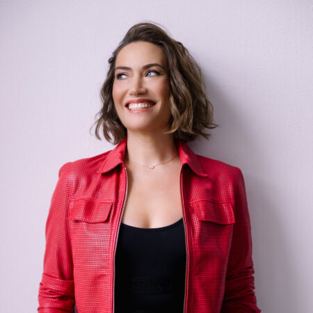 Opera singer Heidi Stober, a smiling woman with wavy, shoulder-length brown hair, wears a textured red jacket over a black top, standing against a light purple wall.