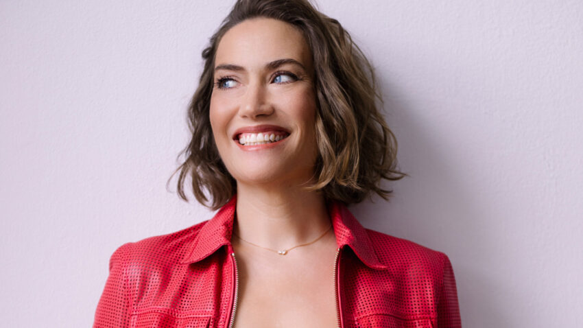 Opera singer Heidi Stober, a smiling woman with wavy, shoulder-length brown hair, wears a textured red jacket over a black top, standing against a light purple wall.