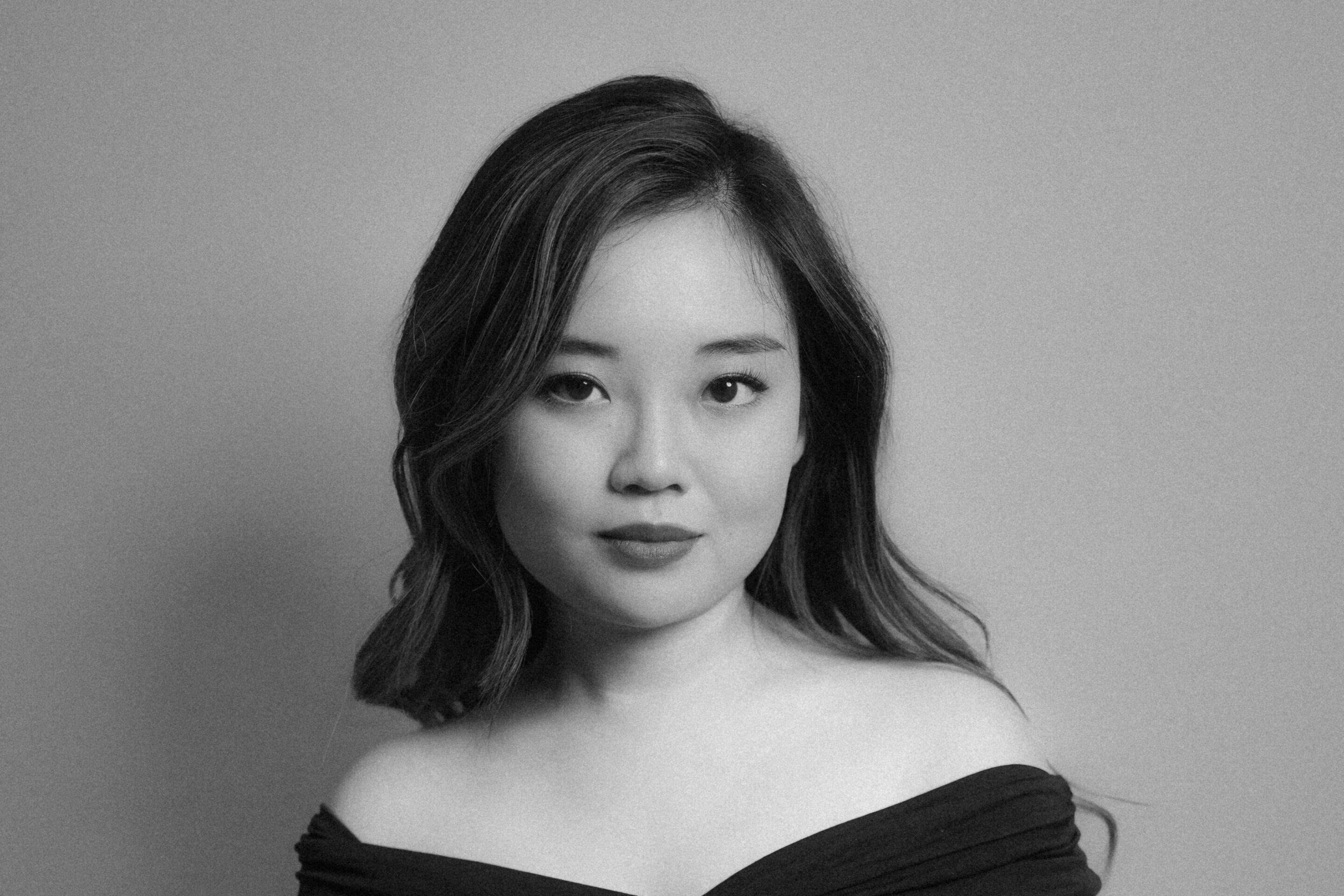 A young woman with long hair poses indoors against a plain background, wearing an off-the-shoulder, dark dress. The photo is black and white, and she gazes confidently at the camera.