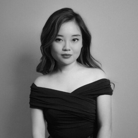 A young woman with long hair poses indoors against a plain background, wearing an off-the-shoulder, dark dress. The photo is black and white, and she gazes confidently at the camera.