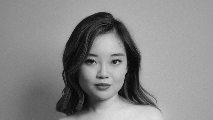 A young woman with long hair poses indoors against a plain background, wearing an off-the-shoulder, dark dress. The photo is black and white, and she gazes confidently at the camera.