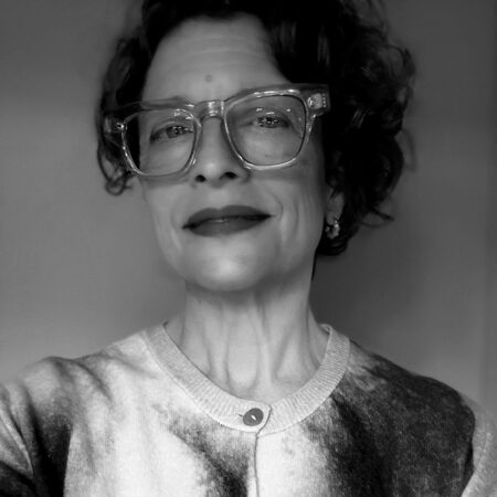 Black and white portrait of Laura Hopkins, a person with short curly hair, wearing large clear-framed glasses and a patterned buttoned shirt, smiling slightly and looking confidently at the camera.
