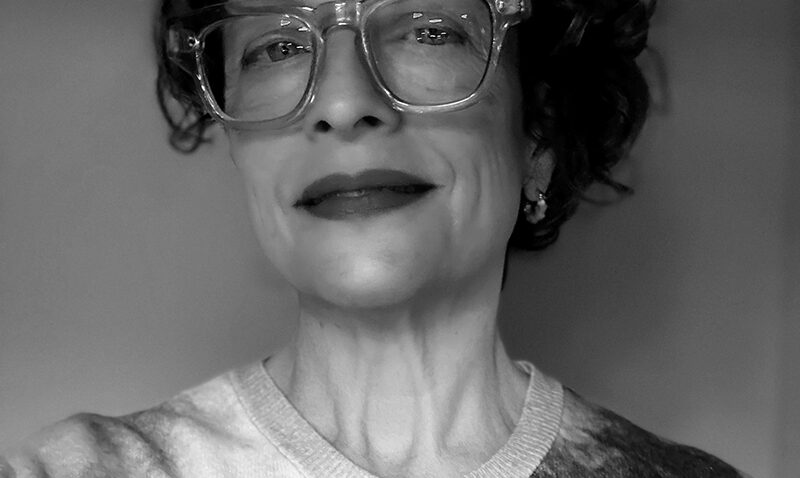 Black and white portrait of Laura Hopkins, a person with short curly hair, wearing large clear-framed glasses and a patterned buttoned shirt, smiling slightly and looking confidently at the camera.