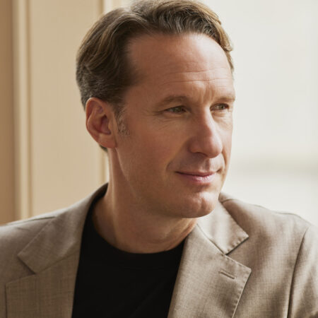 Opera singer Jacques Imbrailo, a renowned baritone with short light brown hair, wearing a beige blazer and black shirt, looks thoughtfully out of a window in soft natural light.