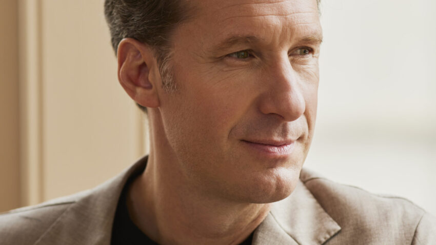 Opera singer Jacques Imbrailo, a renowned baritone with short light brown hair, wearing a beige blazer and black shirt, looks thoughtfully out of a window in soft natural light.