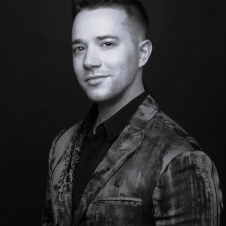 A man with short, styled hair wearing a patterned suit jacket and a dark shirt poses confidently against a dark background. The image is in black and white.