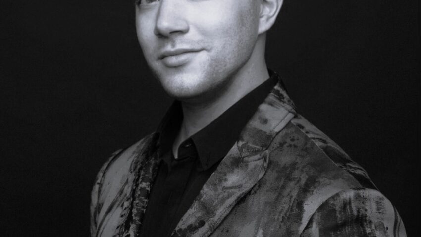A man with short, styled hair wearing a patterned suit jacket and a dark shirt poses confidently against a dark background. The image is in black and white.