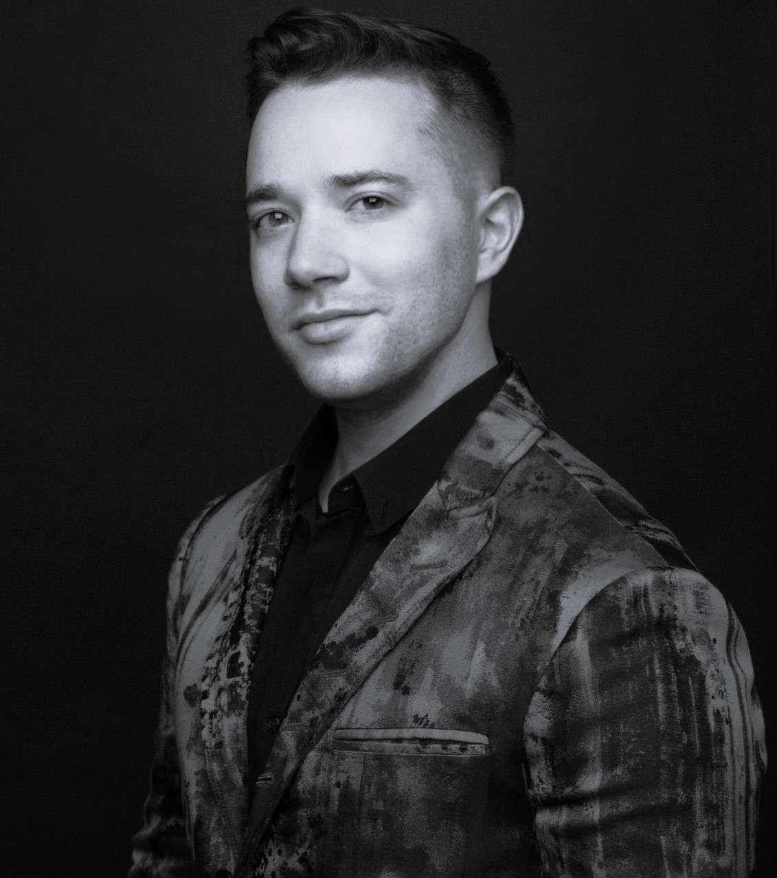 A man with short, styled hair wearing a patterned suit jacket and a dark shirt poses confidently against a dark background. The image is in black and white.