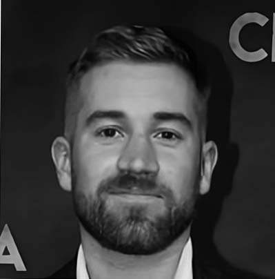 A man with short hair and a beard, possibly Jamie Manton, is smiling slightly whilst facing the camera. The background is dark and partially shows large, cut-off letters.