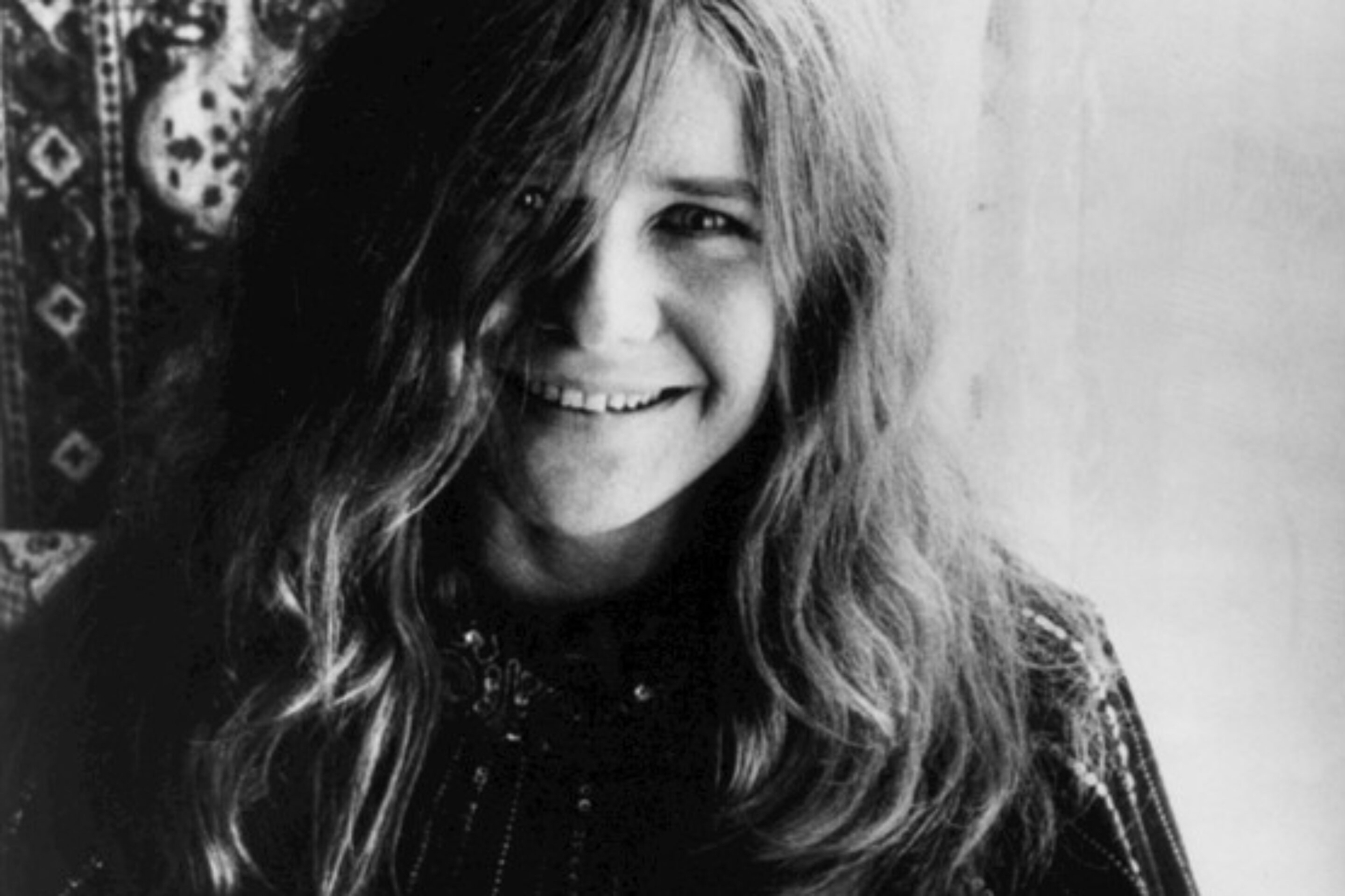 A black-and-white photo of a woman with long hair smiling at the camera, wearing an ornate, patterned garment with swirling designs. She stands before a patterned backdrop, evoking the timeless elegance and history of classic Summertime portraits.