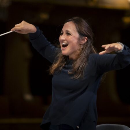 A woman with long brown hair, wearing a dark long-sleeve top, energetically conducts an orchestra with a baton, smiling and gesturing with enthusiasm—just like renowned Portuguese conductor Joana Carneiro—in a dimly lit concert hall.