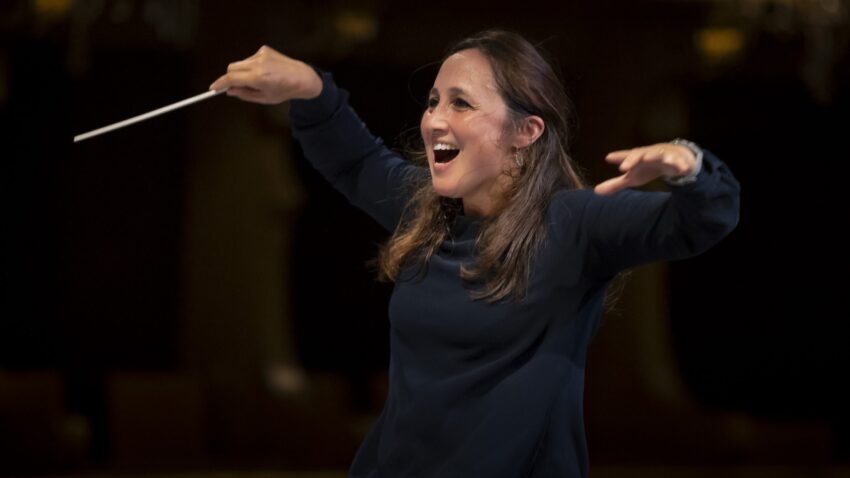 A woman with long brown hair, wearing a dark long-sleeve top, energetically conducts an orchestra with a baton, smiling and gesturing with enthusiasm—just like renowned Portuguese conductor Joana Carneiro—in a dimly lit concert hall.