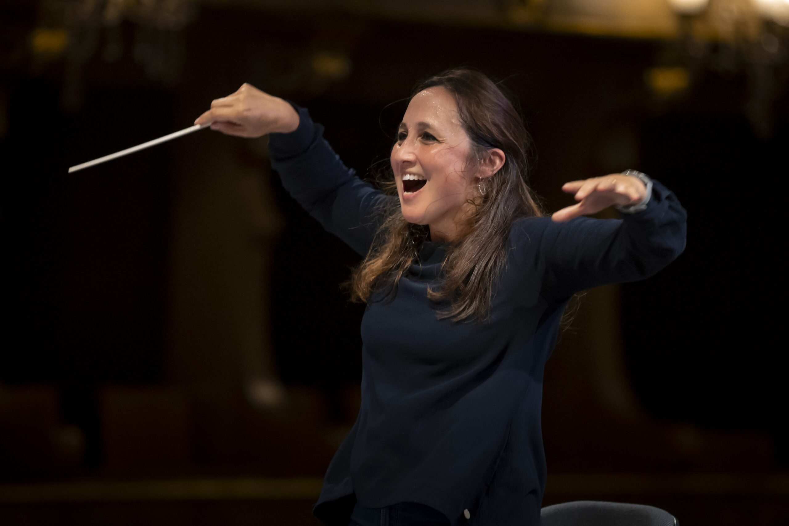 A woman with long brown hair, wearing a dark long-sleeve top, energetically conducts an orchestra with a baton, smiling and gesturing with enthusiasm—just like renowned Portuguese conductor Joana Carneiro—in a dimly lit concert hall.