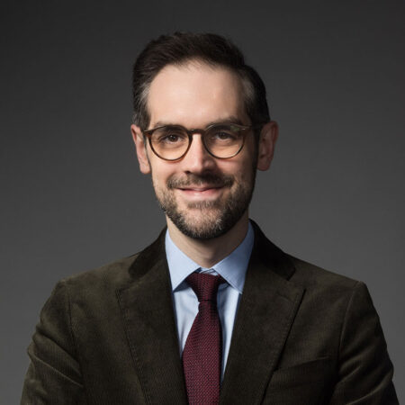 John Savournin, a man with short dark hair, glasses, and a beard, wears a brown suit jacket, blue shirt, and red tie, smiling slightly against a plain dark background.