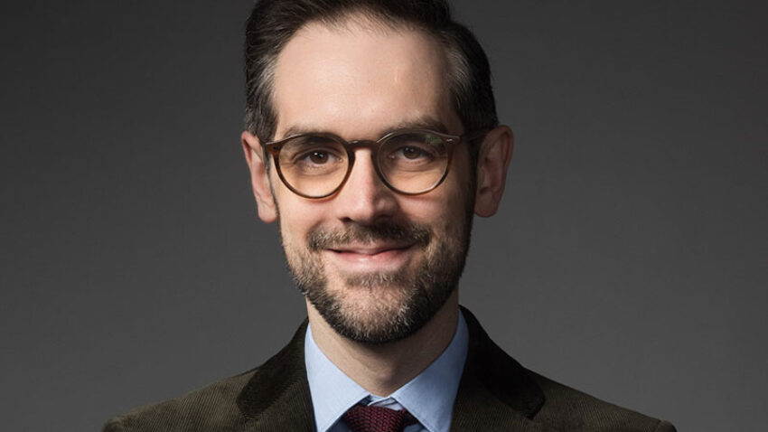 John Savournin, a man with short dark hair, glasses, and a beard, wears a brown suit jacket, blue shirt, and red tie, smiling slightly against a plain dark background.