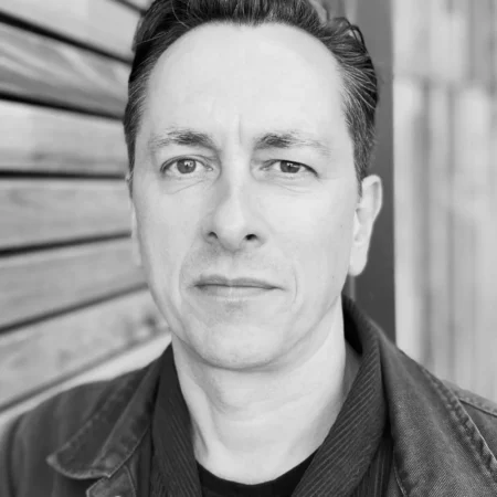 Black and white portrait of a man with short dark hair, wearing a dark jacket and shirt, standing in front of a wooden wall and looking directly at the camera with a neutral expression.