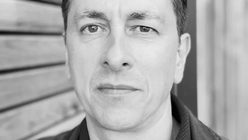 Black and white portrait of a man with short dark hair, wearing a dark jacket and shirt, standing in front of a wooden wall and looking directly at the camera with a neutral expression.