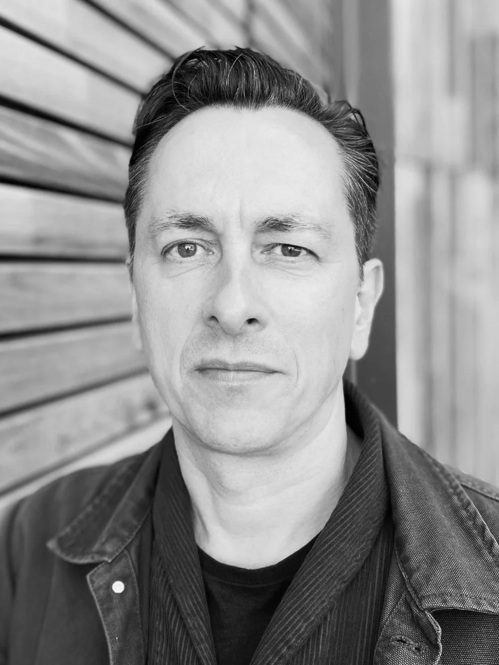 Black and white portrait of a man with short dark hair, wearing a dark jacket and shirt, standing in front of a wooden wall and looking directly at the camera with a neutral expression.