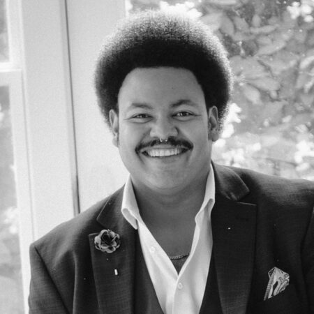 Joshua Blue, a man with an afro hairstyle and moustache smiles warmly at the camera. He is wearing a dark jacket with a buttonhole flower and a pocket square, a white shirt, and a chain necklace. The photo is black and white.