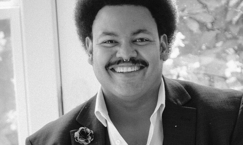 Joshua Blue, a man with an afro hairstyle and moustache smiles warmly at the camera. He is wearing a dark jacket with a buttonhole flower and a pocket square, a white shirt, and a chain necklace. The photo is black and white.
