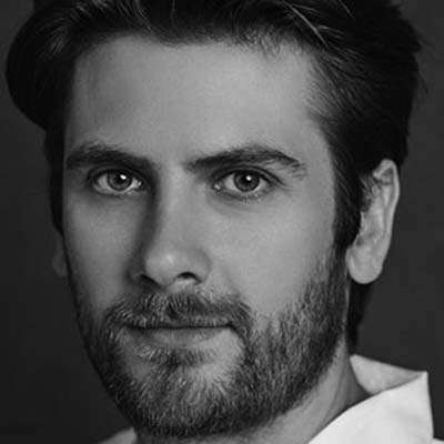 Black and white close-up portrait of a man with dark hair and a beard, looking directly at the camera with a neutral expression, wearing a light-coloured shirt.
