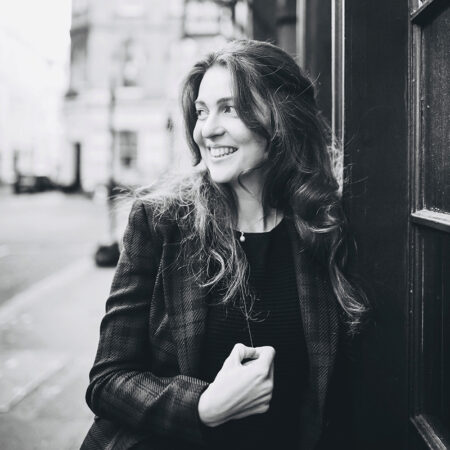 Black and white photo of Julia Burbach, a smiling woman with long hair, wearing a blazer and leaning against a building on a city street. She looks to the side with her hand near her chest, as the background remains slightly blurred.