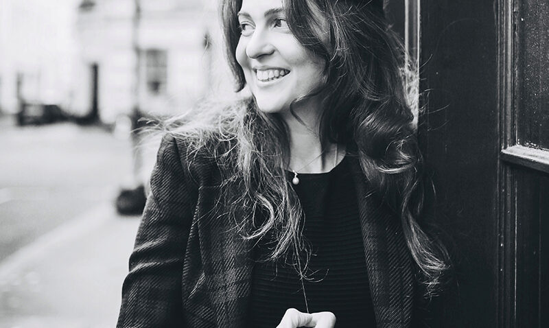 Black and white photo of Julia Burbach, a smiling woman with long hair, wearing a blazer and leaning against a building on a city street. She looks to the side with her hand near her chest, as the background remains slightly blurred.