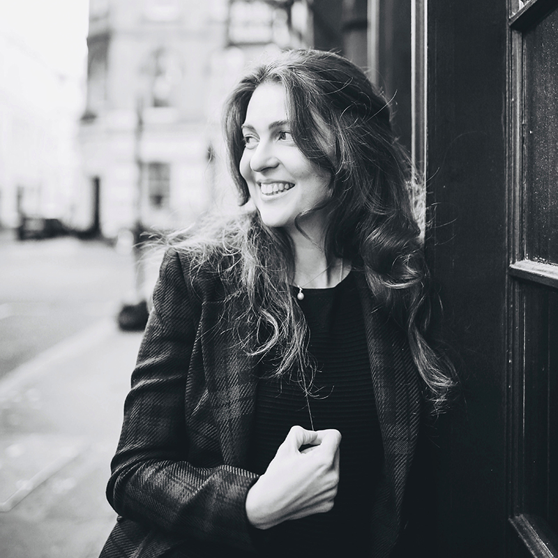 Black and white photo of Julia Burbach, a smiling woman with long hair, wearing a blazer and leaning against a building on a city street. She looks to the side with her hand near her chest, as the background remains slightly blurred.
