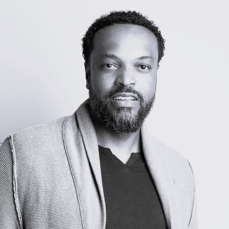Kenneth Kellogg, a man with short curly hair and a beard, wearing a textured jacket over a dark shirt, stands against a plain light background and looks at the camera with a slight smile.