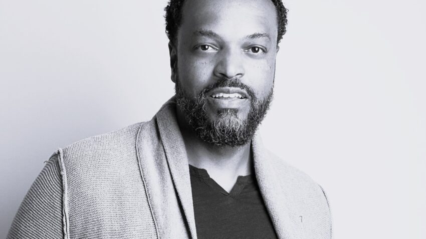 Kenneth Kellogg, a man with short curly hair and a beard, wearing a textured jacket over a dark shirt, stands against a plain light background and looks at the camera with a slight smile.