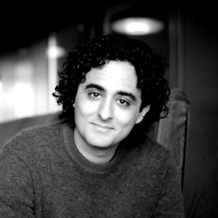 A person with curly dark hair and a gentle smile wears a jumper and sits on a chair in an indoor setting. The image is black and white, with a softly blurred background.