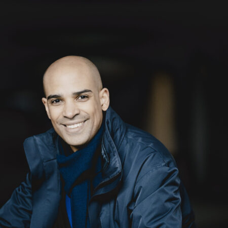 A smiling Kevin John Edusei with a shaven head wears a dark blue jacket and scarf. He is seated and facing the camera, with a dark, blurred background.