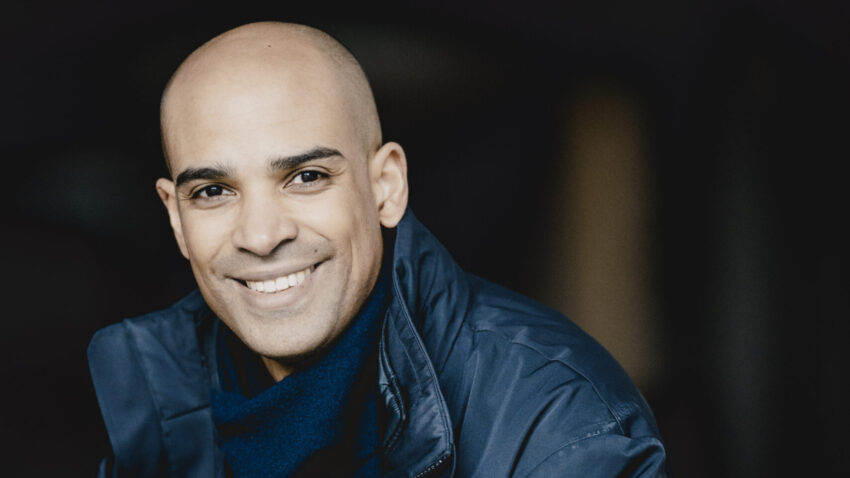 A smiling Kevin John Edusei with a shaven head wears a dark blue jacket and scarf. He is seated and facing the camera, with a dark, blurred background.